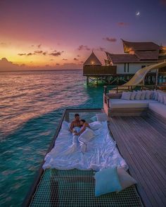 a man laying on top of a bed next to the ocean at sunset in front of an over water villa