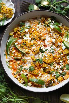 a white bowl filled with mexican food and garnished with cilantro, limes, corn, and feta cheese