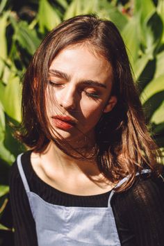 a woman with her eyes closed standing in front of some plants and looking off to the side