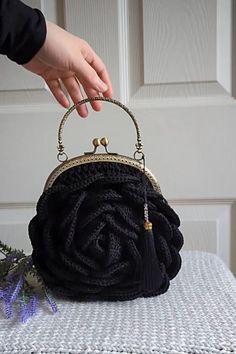 a hand holding a black purse on top of a white table next to a purple flower