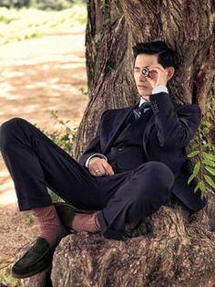 a man in a suit and tie sitting next to a tree talking on a cell phone