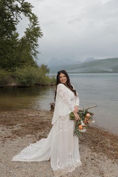 a woman in a wedding dress standing on the shore of a lake with her bouquet