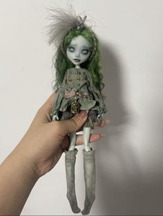 a hand holding a doll with green hair and blue eyes, wearing a gray dress