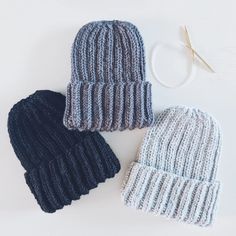 two knitted hats sitting next to each other on a white surface with knitting needles