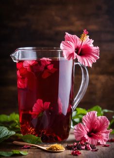 a glass pitcher filled with red liquid and pink flowers next to green leaves on a wooden table