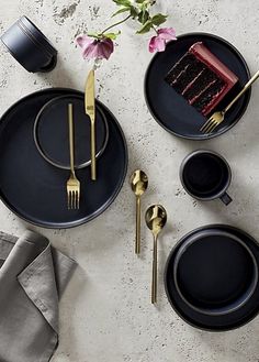 a table with black plates, gold forks and silverware on it next to a pink flower