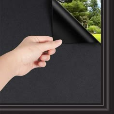 a hand holding a black piece of paper in front of a window with trees and grass