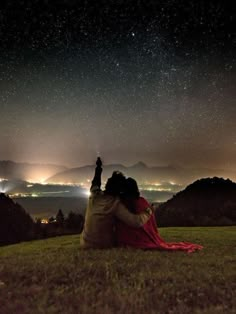 two people sitting on top of a grass covered hill under a night sky filled with stars