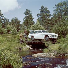 a white car is crossing a bridge over a stream in the woods with people standing on it