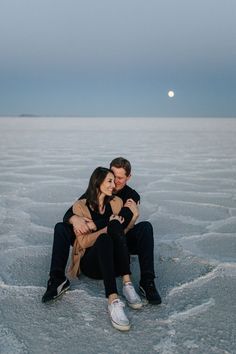 a man and woman sitting in the middle of an icy lake at night with the moon behind them