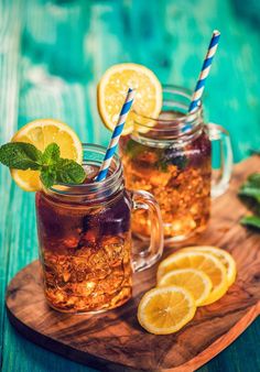 two mason jars filled with iced tea and lemon slices on a cutting board next to mint leaves