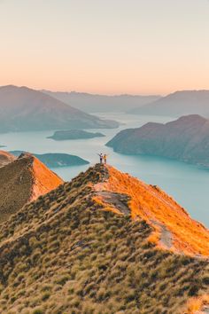 two people standing on the top of a mountain overlooking water and mountains in the distance