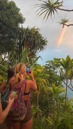 three women are standing in front of some trees and one is taking a photo with her cell phone