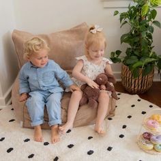 two young children sitting on a chair with stuffed animals in front of them and a potted plant behind them