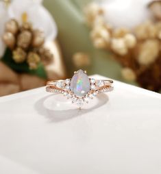 a white opal and diamond ring sitting on top of a table with flowers in the background