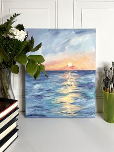 a painting of the sun setting over the ocean on canvas next to some books and flowers