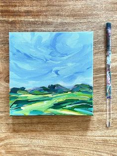 an acrylic painting of green hills and blue sky on a wooden table with a pen