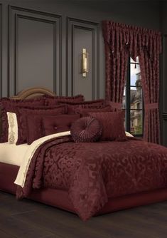 a bed with red comforter and pillows in a room next to a window filled with curtains