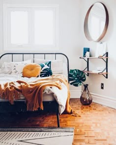 a bedroom with white walls and wood flooring has a metal bed frame, round mirror, and plant on the wall