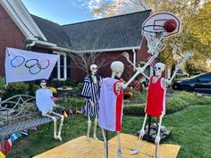 three skeleton figurines in front of a house with an olympic flag and basketball hoop