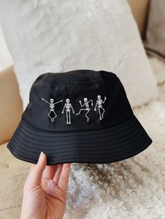 "Complete your trendy look with this Cara Mak Halloween Bucket Hat. This 100% cotton twill bucket hat comes in a solid black colorway and includes embroidered dancing skeletons at the crown.  ☆ Size: 3 ¾″ (7.6 cm) crown, 2 ¼″ (5.1 cm) brim & 22\" (55.88cm) circumference ☆ Embroidered Skeletons at Crown ☆ Stiff Brim ☆ 100% Cotton Twill ☆ One Size Fits Most ☆ Sewn Eyelets for Breathability 🌈 I SHIP ALL HATS WITHIN 1-2 WEEKS ☆ All items are shipped via USPS  ☆ Items take 3-5 business days to deliver after it is dropped off at the post office (EDIT: shipping times may vary due to the recent outbreak of COVID-19) 🍄 JOIN THE EMAIL LIST FOR 15% OFF YOUR NEXT ORDER: ☆ Make sure you're on my email list to get access to exclusive & private sales & products! ☆ http://caramak.com/email-signup 🍄 Per Black Novelty Hat For Halloween, Adjustable Skull Print Hats For Halloween, Adjustable Skull Print Halloween Hats, Adjustable Cotton Hat For Halloween, Adjustable Cotton Halloween Hat, Black Cotton Halloween Hat, Halloween Cotton Hat, Black Novelty Cotton Hat, Fun Black Halloween Hat