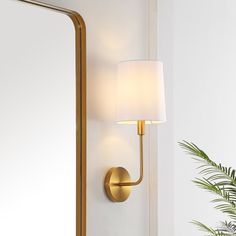 a wall light with a white shade on it next to a mirror and potted plant