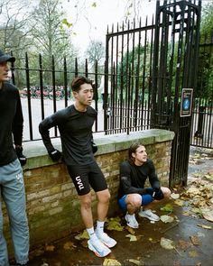 two men standing next to each other near a brick wall and iron fence with leaves on the ground