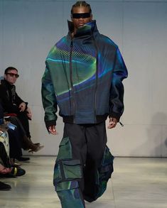 a man walking down a runway in a blue jacket and black pants with an abstract print on it