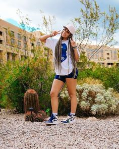 a woman with dreadlocks is standing in front of some bushes and cactuses