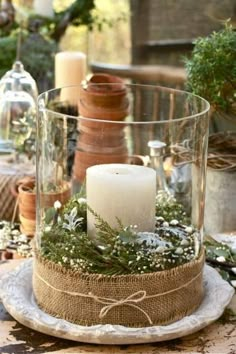 a table topped with a glass vase filled with candles and greenery next to potted plants