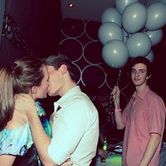 two people standing next to each other with balloons in the air behind them and one person kissing another man