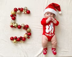 a baby in a santa hat laying next to the letter e on a bed with christmas decorations