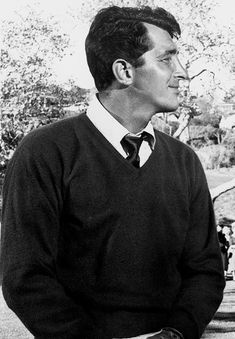 a man in a sweater and tie sitting on a park bench looking off into the distance