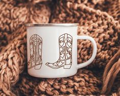 a white coffee cup sitting on top of a pile of brown knitted blankets next to a pair of boots