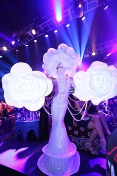 a woman in white dress standing on stage with lights behind her and flowers around her