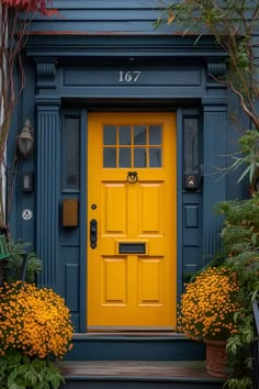 a yellow door is in front of a blue house with flowers on the steps and bushes around it