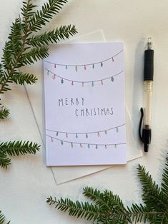 a christmas card next to a pen on top of a white surface with greenery