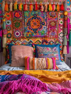 a bed covered in lots of colorful pillows next to a wall hanging with tassels