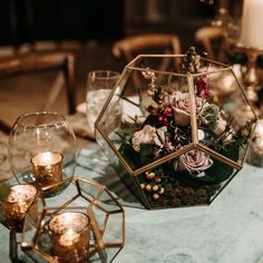 a table topped with glass vases filled with flowers and greenery next to candles