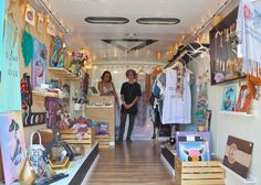 two women standing in the doorway of a store with lots of items on shelves and clothes hanging from the ceiling