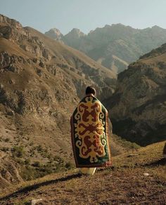 a man sitting on top of a hill covered in a blanket and looking at the mountains