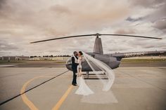 a bride and groom standing in front of a helicopter on the tarmac at an airport