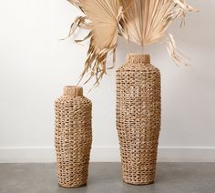 two tall vases with plants in them sitting on a cement floor next to a white wall