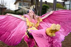 a woman in pink and yellow costume swinging on a swing with her wings spread out