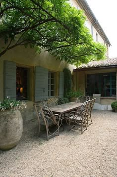 an outdoor table and chairs in front of a house with large potted plants on the patio