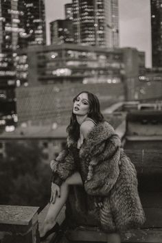 black and white photograph of woman in fur coat sitting on ledge with city skyline in background