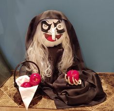 a creepy doll with long white hair wearing a black cape and holding two red apples