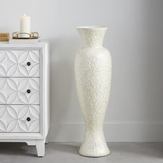 a tall white vase sitting next to a dresser with a candle on top of it