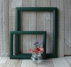 two square frames with flowers in them against a wooden wall behind one is a glass vase