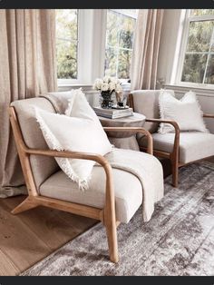 a living room with two chairs and a table in front of a window that has curtains on the windowsill